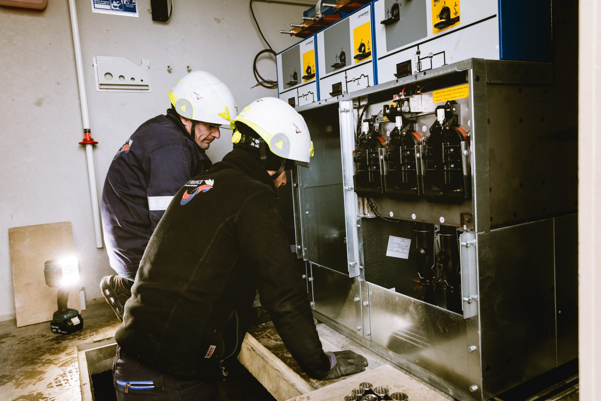 Deux techniciens casqués interviennent sur des cellules haute tension ouvertes, dans un poste électrique